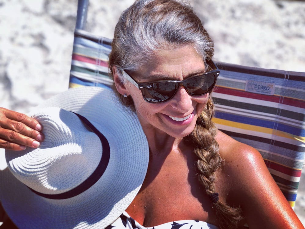 image of woman with long braid and gray roots wearing sunglasses sitting on a beach chair holding a hat