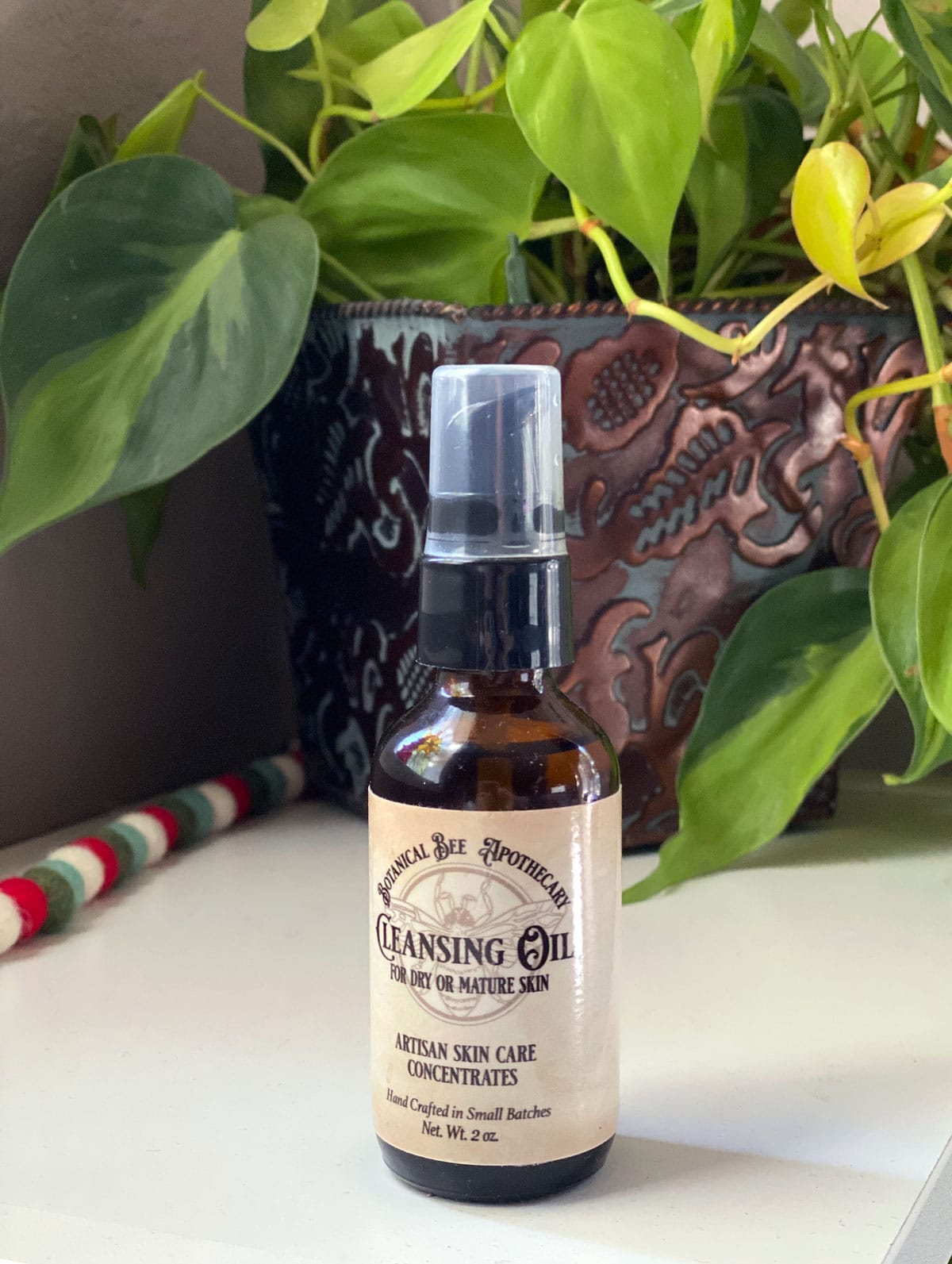 bottle of Botanical Bee cleansing oil on a white counter next to a plant and a Christmas garland